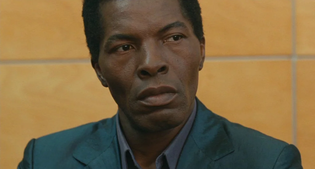 The Limits of Control (2009) movie still: closeup — A man stands before a wall covered in light-colored square wood tiles. He gazes steadily towards…