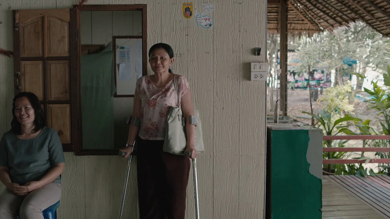 Cemetery of Splendour (2015) movie still: fullbody — A light wood-paneled wall sits next to an open, thatched-roof porch area. One woman stands…