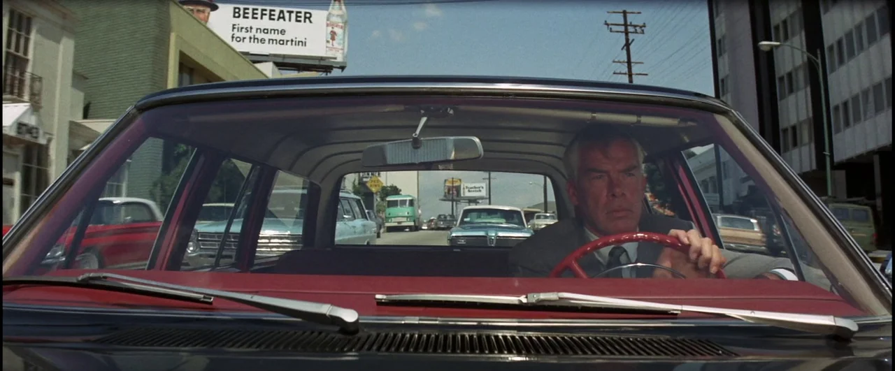 Point Blank (1967) movie still: medium — A man drives a car through a mid-century city street lined with billboards. The man keeps both hands on the…