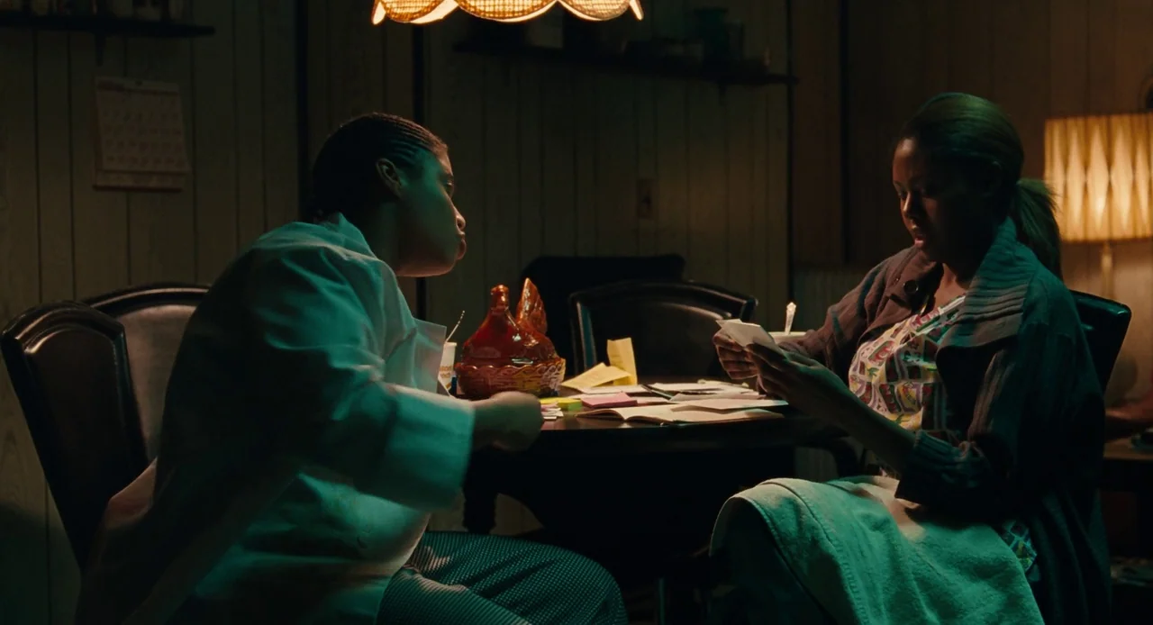 Pariah (2011) movie still: medium — A dimly lit room with wood-paneled walls and retro-style decor at night. Two individuals sit at a small round…