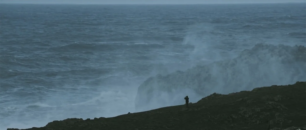 Taking Back the Waves (2017) movie still: wide — A rugged, rocky coastline overlooks a turbulent sea with heavy crashing waves. A lone person stands…