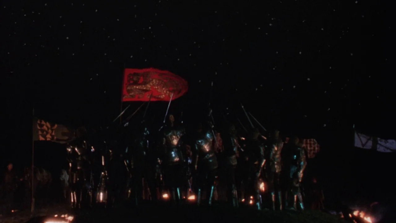 Excalibur (1981) movie still: wide — A group of armored knights stands on a dark hill at night. They hold spears and banners beneath a clear sky…