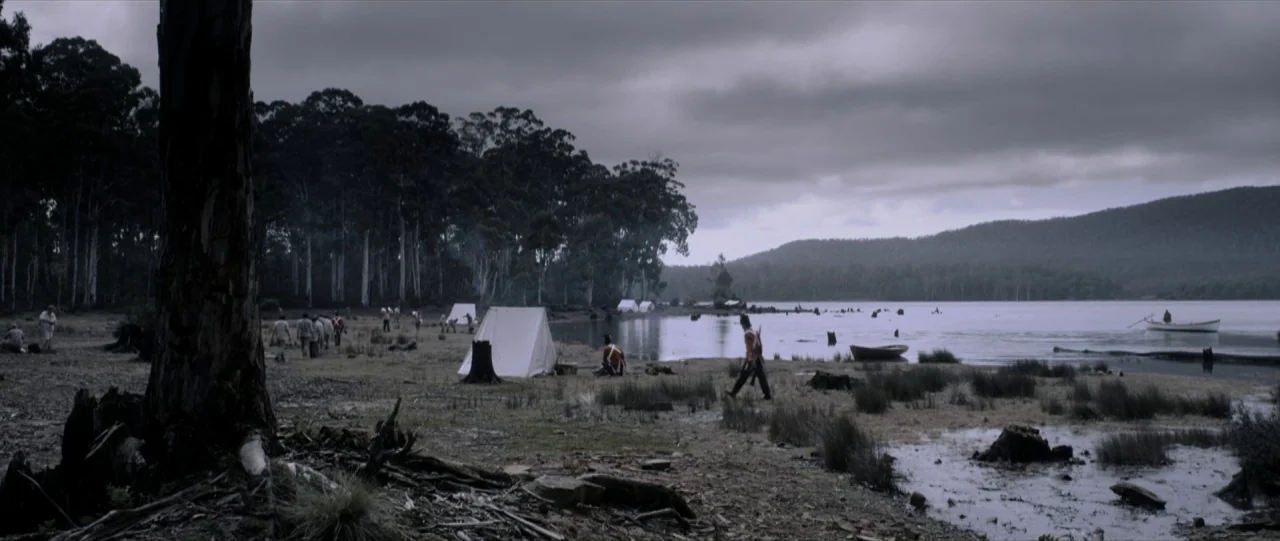 Van Diemens Land (2009) movie still: wide — A muddy shoreline sits beside a large lake under a heavy, overcast grey sky. Several white canvas tents…