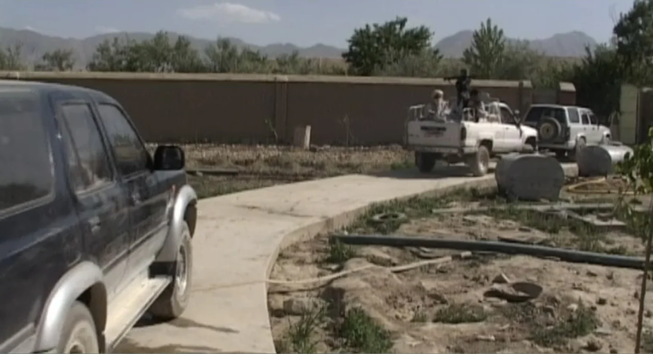 Afghanistan's Opium Trail (2007) movie still: wide — A dry, sunlit dirt lot features a concrete walkway bordered by a mud wall. Men ride in the bed…