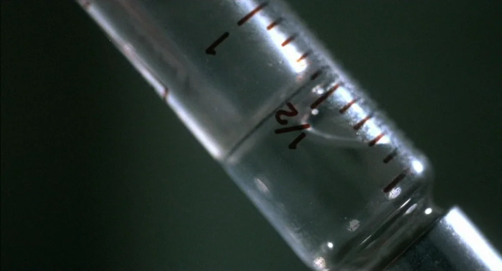 Drugstore Cowboy (1989) movie still: closeup — A close-up view shows a glass syringe with measurement markings against a dark backdrop. The syringe…
