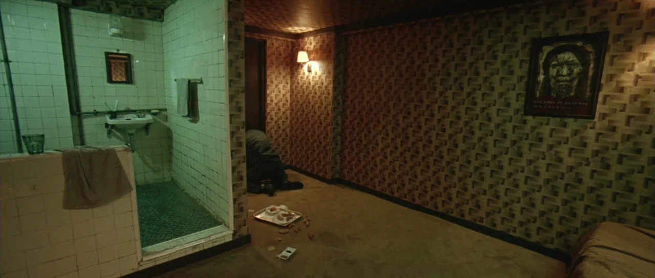 Oldboy (2003) movie still: wide — A small motel room features tiled bathroom walls painted green and textured brown wallpaper. A man in dark…