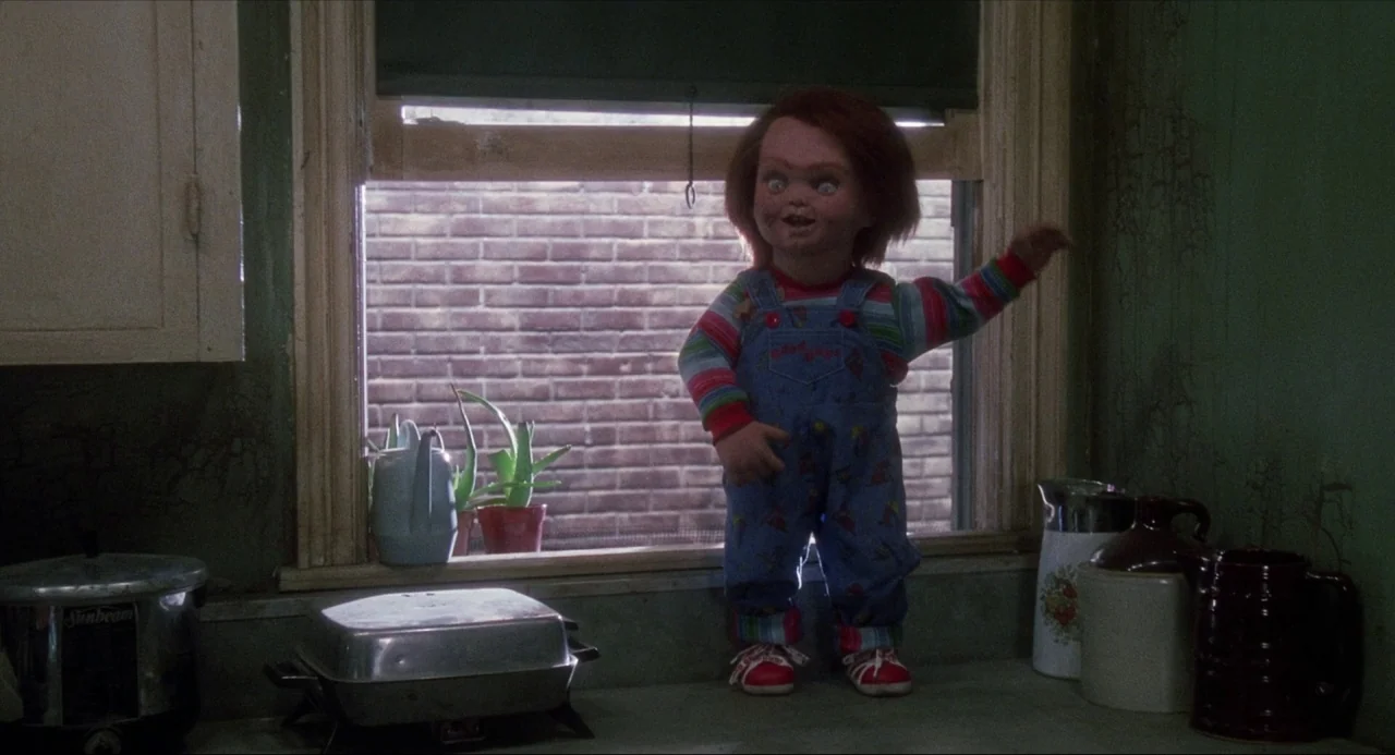 Child’s Play (1988) movie still: medium — A dark, cluttered kitchen features a doll standing on a surface near a window. The doll holds one arm up…