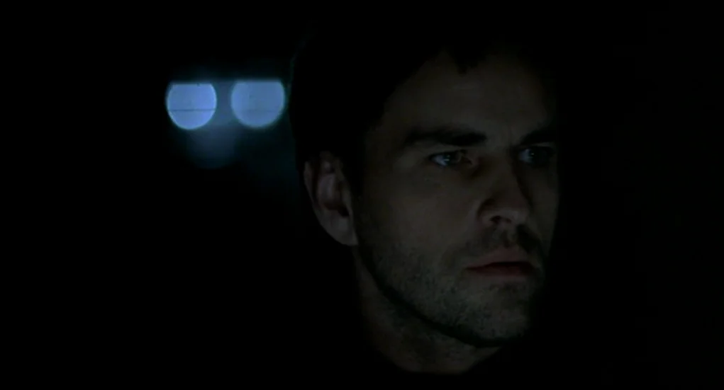 Tiresia (2003) movie still: closeup — A dark car interior at night with a blurred, glowing light in the background. A man sits in the driver's seat…
