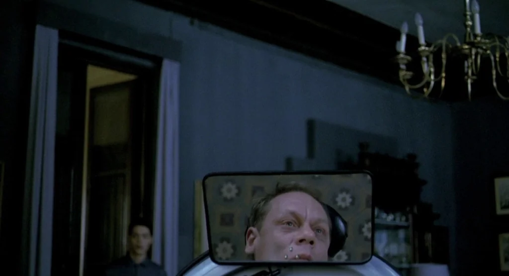 In a Glass Cage (1986) movie still: medium — A dimly lit room features a man reflected in a mirror. A second person stands blurred in the dark…
