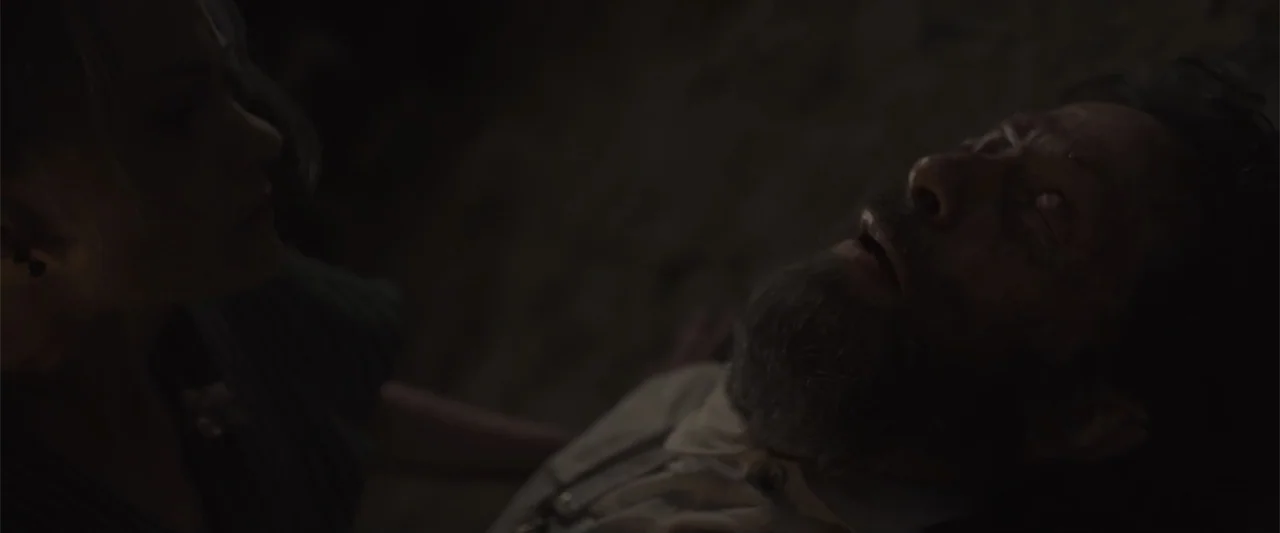 Ghosts of the Ozarks (2022) movie still: closeup — A dark indoor space where a man lies on the floor. A woman stands over him while looking down at…