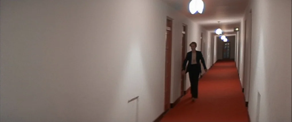 The Man Who Fell To Earth (1976) movie still: fullbody — A long hotel corridor features stark white walls and a bright red carpet runner. A man in a…
