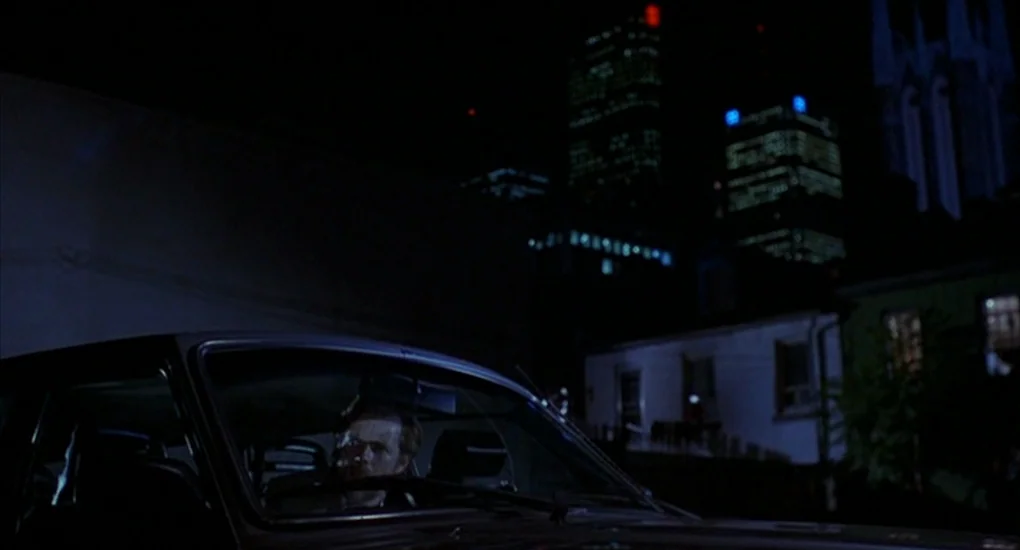 Exotica (1994) movie still: medium — A man sits inside a car parked on a dark urban street at night. Tall city buildings with illuminated windows…