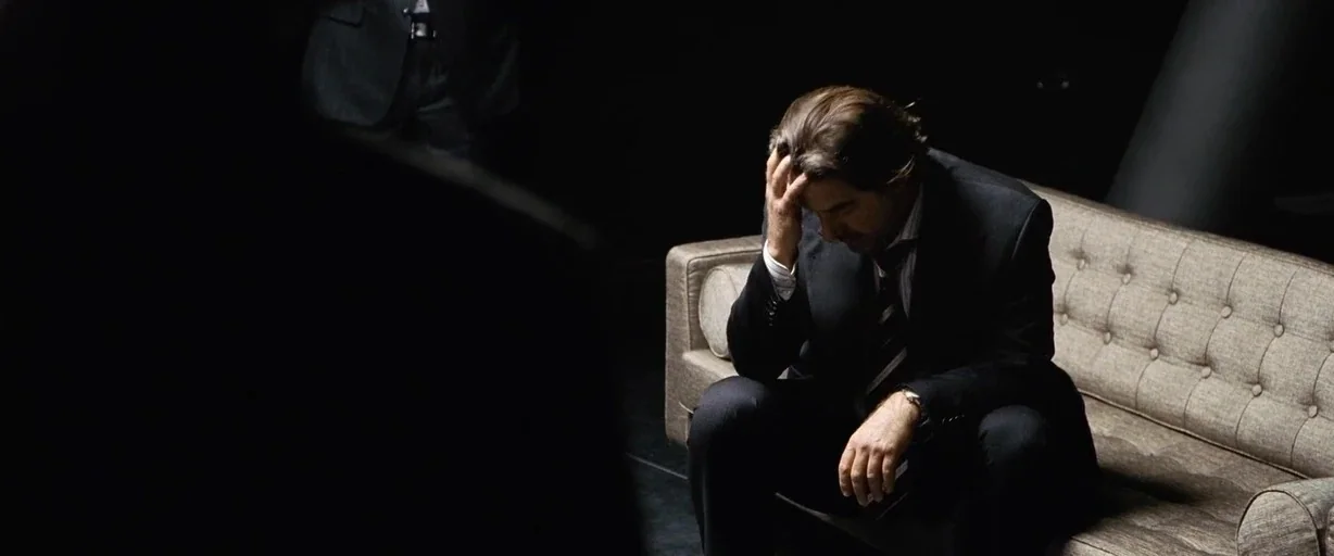 The International (2009) movie still: medium — A dimly lit room features a tufted fabric sofa positioned against a dark wall. A man in a suit sits…