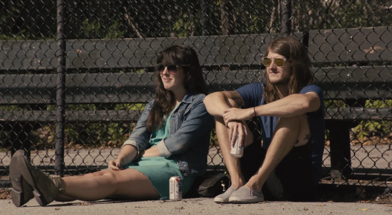 The Comedy (2012) movie still: fullbody — A pair of young adults sit on a paved surface against a chain link fence. They are relaxing next to a…