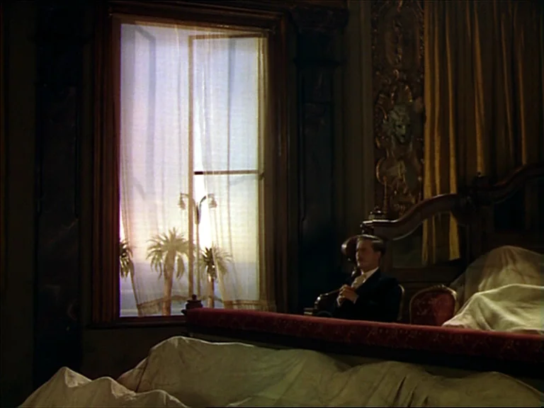 The Red Shoes (1948) movie still: wide — A dimly lit, ornate room features a large window with sheer white curtains. A man in a dark suit sits on…