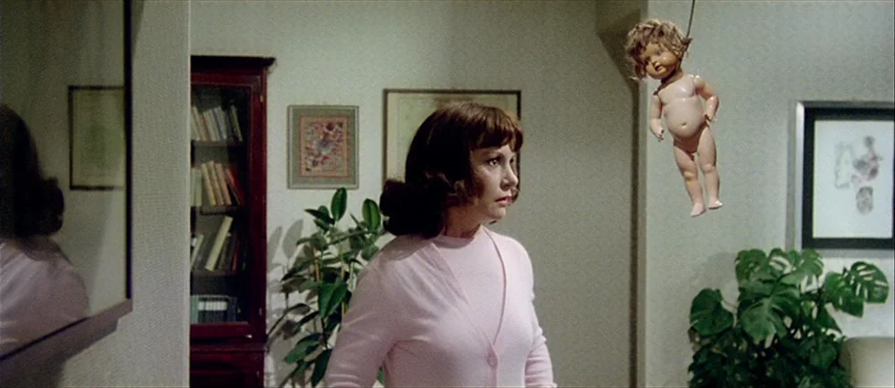 Deep Red (1975) movie still: medium — A living room interior featuring a dark wooden bookcase and decorative framed wall art. A woman stands in the…