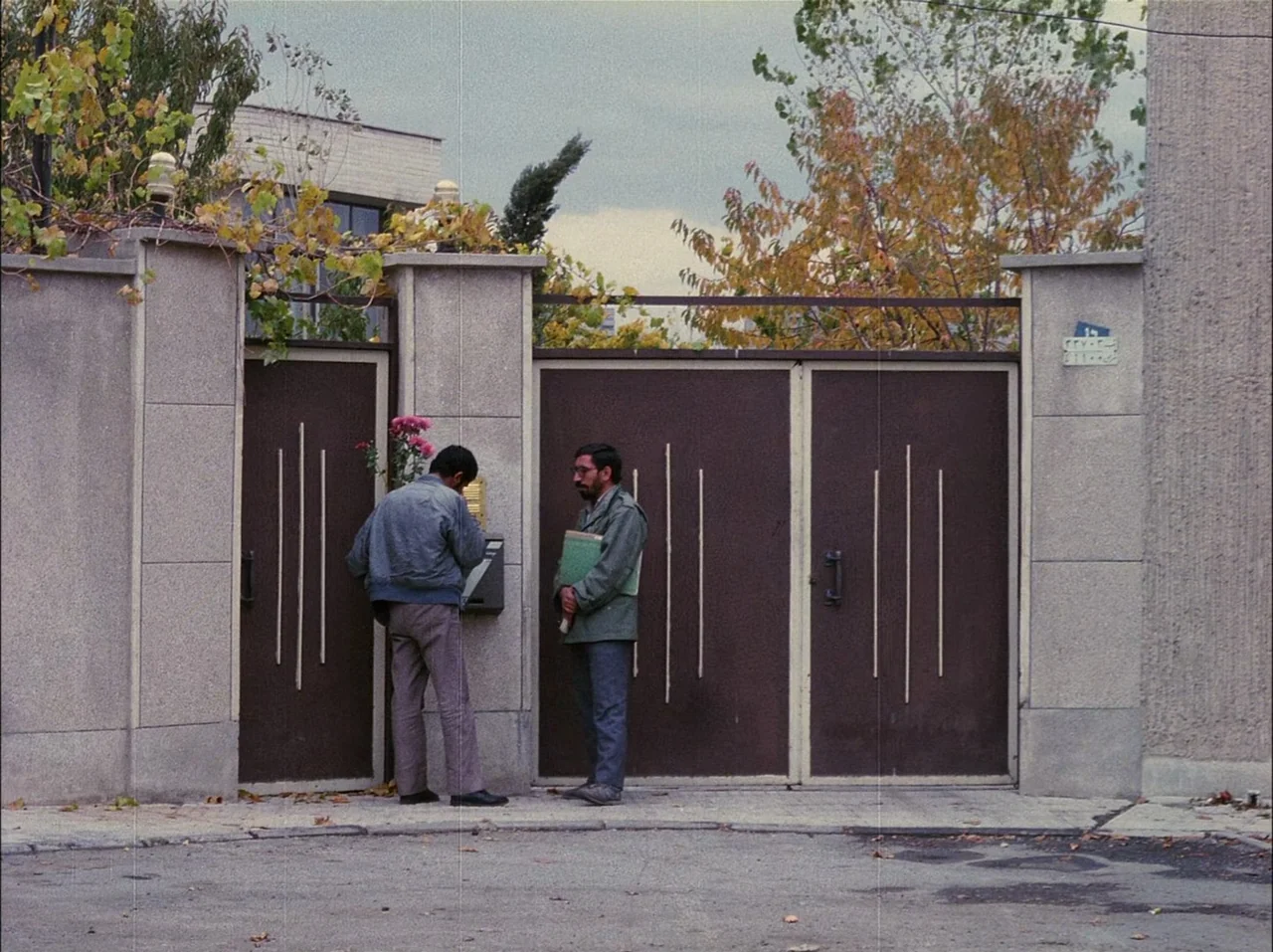 Close-Up (1990) movie still: fullbody — A residential entrance with a brown metal gate set into a concrete wall. Two men stand together outside the…