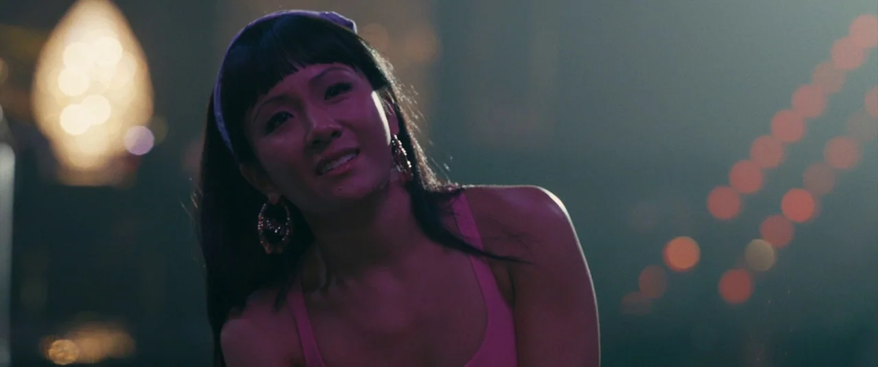 Hustlers (2019) movie still: medium — A woman stands on a dimly lit city street at night with blurry background lights. She wears a headscarf and a…