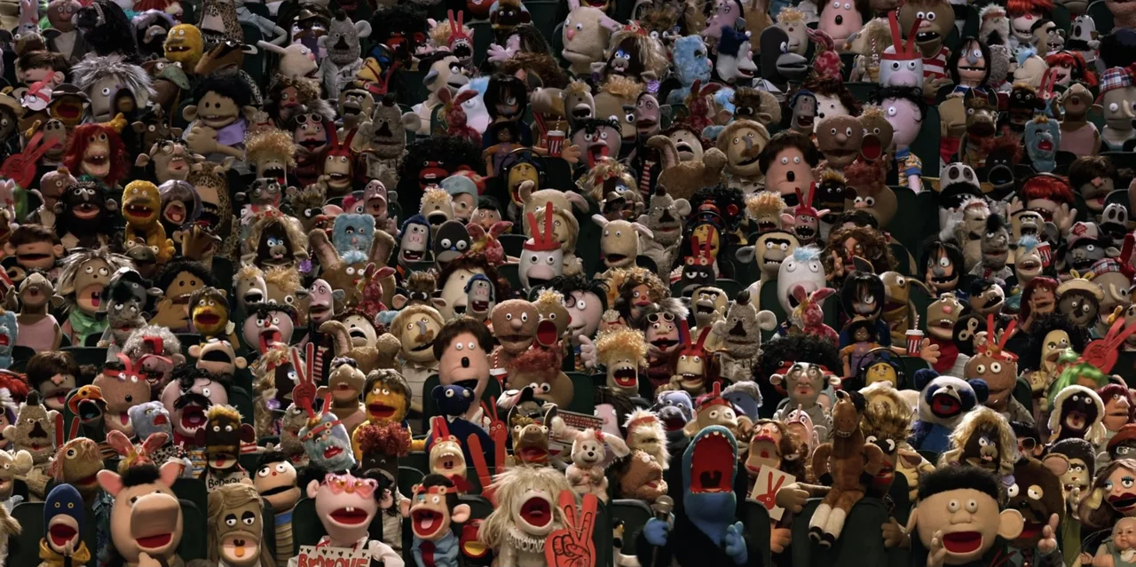 31 Minutes: One Hot Christmas (2025) movie still: wide — A dense crowd of various fabric puppets fills the seats of a large theater. The puppets…