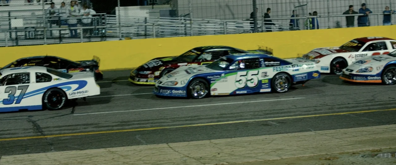 God Bless the Broken Road (2018) movie still: wide — A paved asphalt racetrack features multiple stock cars driving during a night event. Spectators…