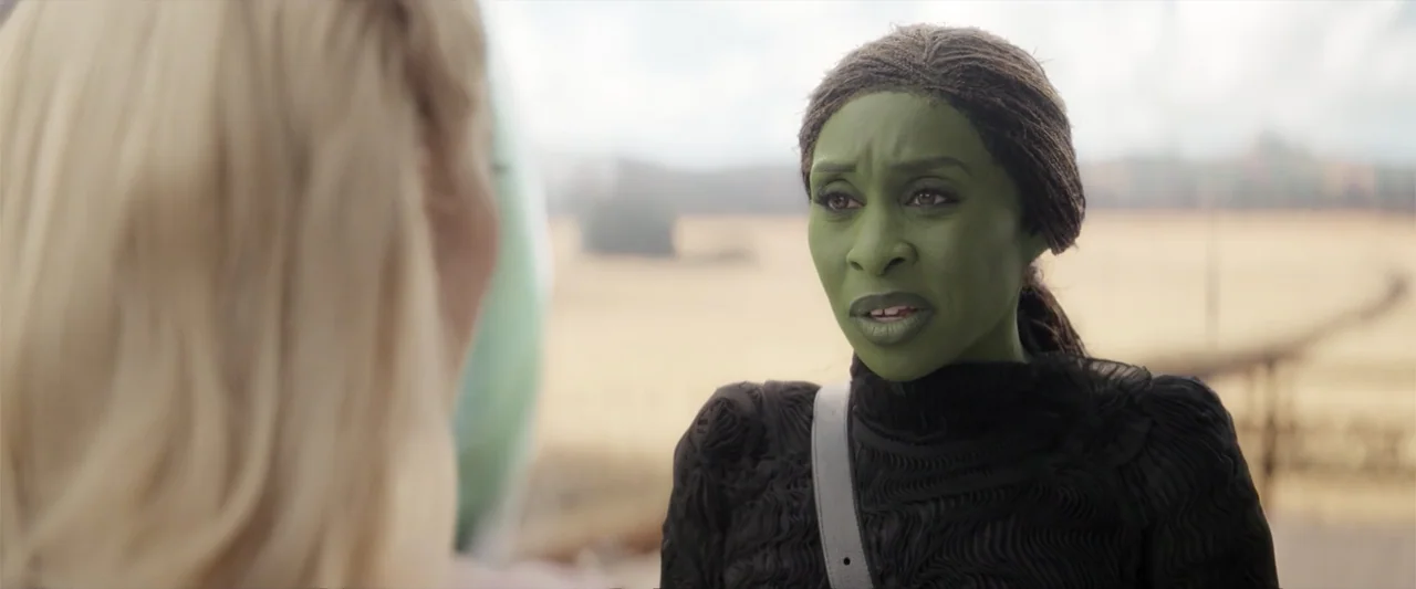 Wicked (2024) movie still: medium — A field with dry grass serves as the backdrop under a cloudy sky. A woman with green skin wears a black textured…
