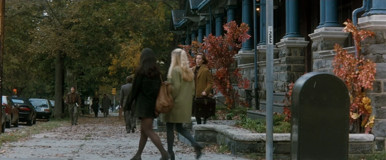 Fallen (2004) movie still: medium — A tree-lined sidewalk sits beside a stone building with blue porch pillars in autumn. Two women walk away from…