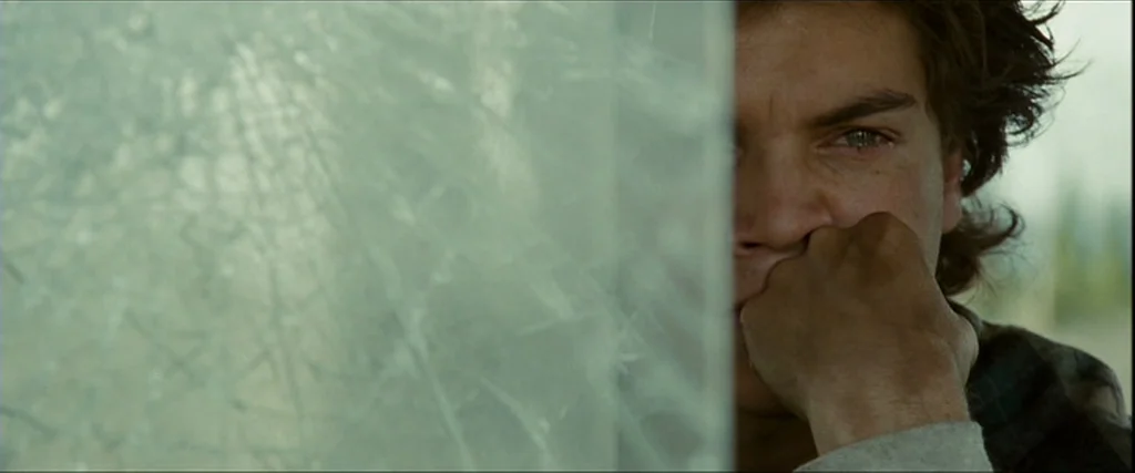 Into the Wild (2007) movie still: closeup — A man's face is partially obscured by a cracked, semi-transparent pane of glass. He rests his hand…