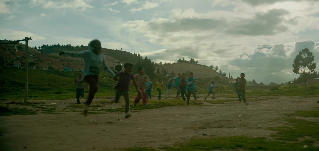 Triple Frontier (2019) movie still: wide — An open dirt field stretches out before a backdrop of grassy hills and sparse houses. A group of children…