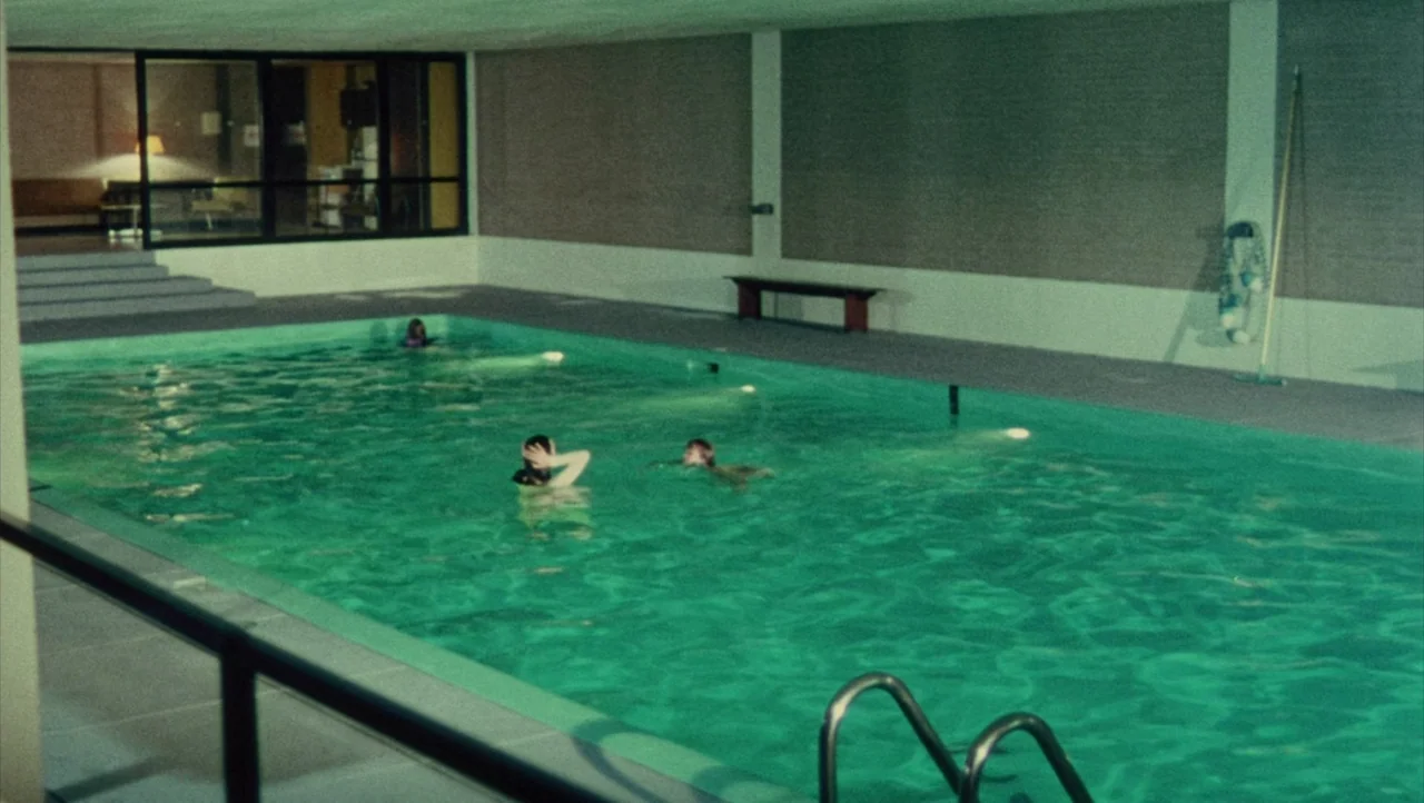 Shivers (1975) movie still: wide — A spacious indoor swimming pool room features smooth concrete walls and a glass-enclosed viewing area. Three…
