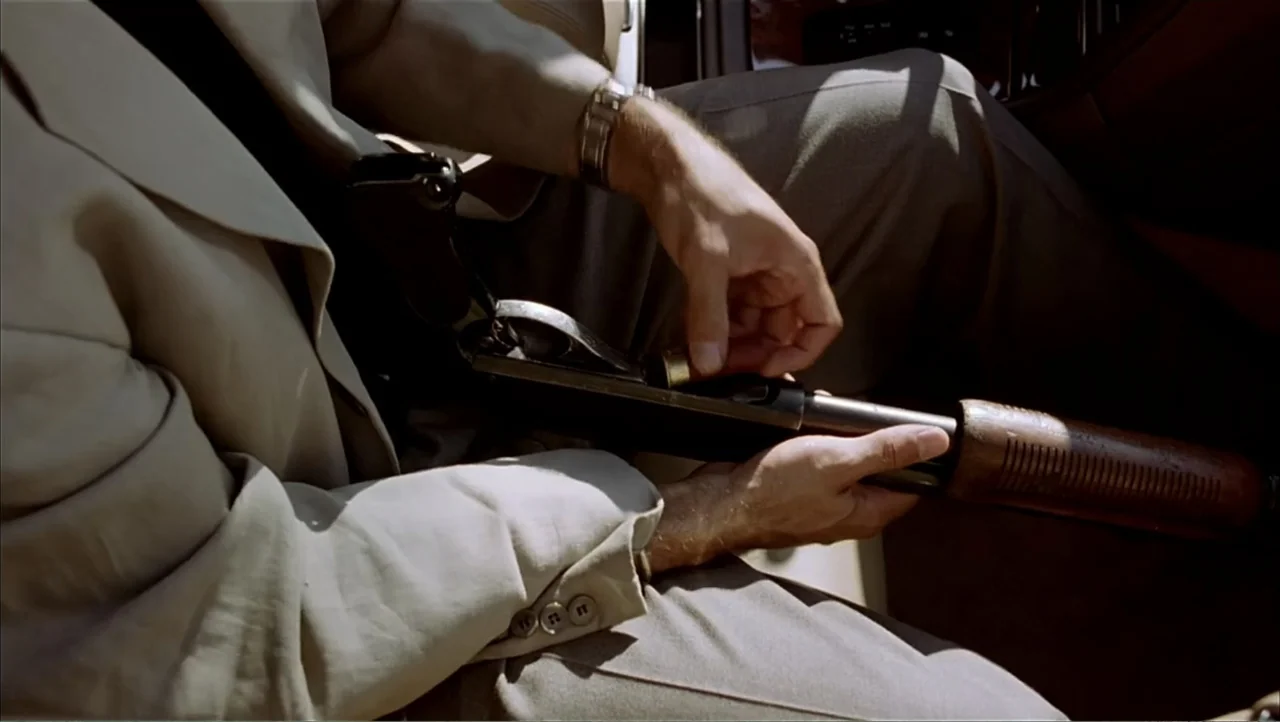 Ajuste de Cuentas (2016) movie still: closeup — Inside a car interior, a person wears a tan suit jacket. Hands load a single shell into the chamber…