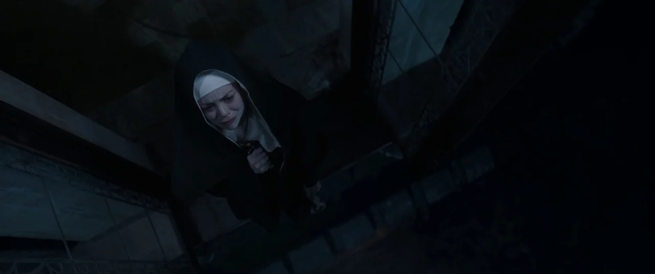 The Nun (2018) movie still: medium — A dark, stone exterior stairwell at night with minimal illumination from above. A young nun in a habit stands…