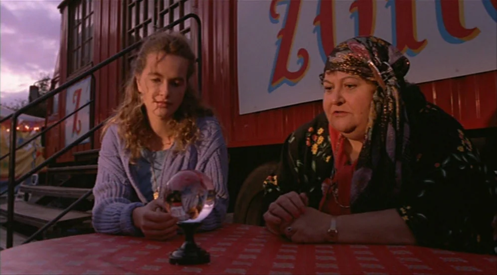 Roselyne and the Lions (1989) movie still: medium — A red carnival wagon stands in an outdoor area during the late afternoon. Two women sit at a red…