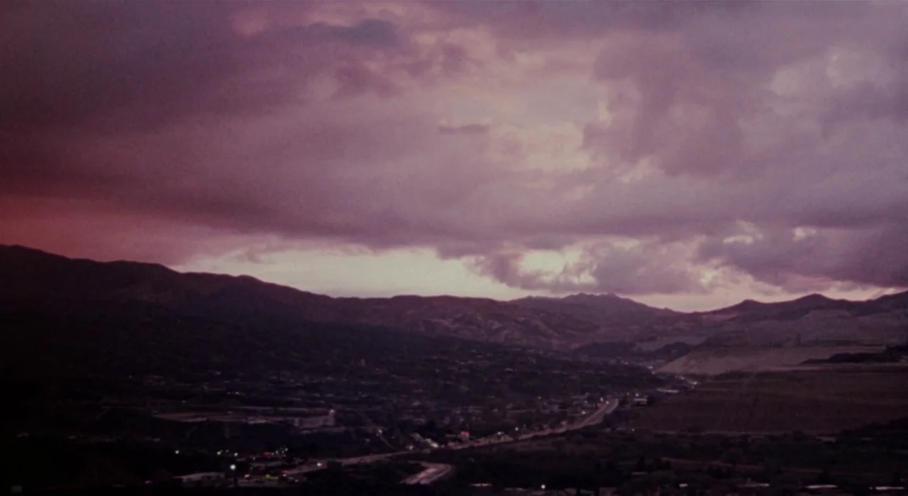 White Of The Eye (1987) movie still: establishing — A vast mountain valley sits beneath a heavy, purple-tinted cloud cover at dusk. Sparse lights…