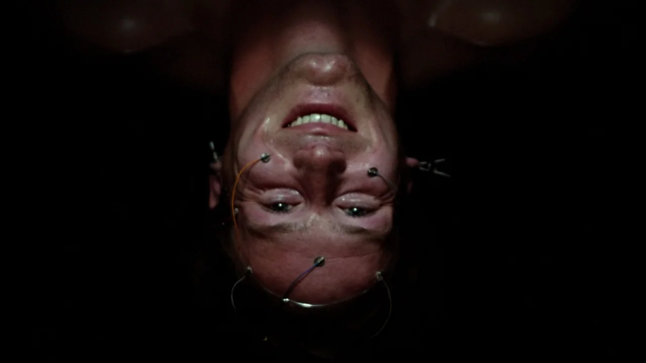 Altered States (1980) movie still: closeup — A man is positioned upside down in a pitch-black, indeterminate space. Wires with small metal…
