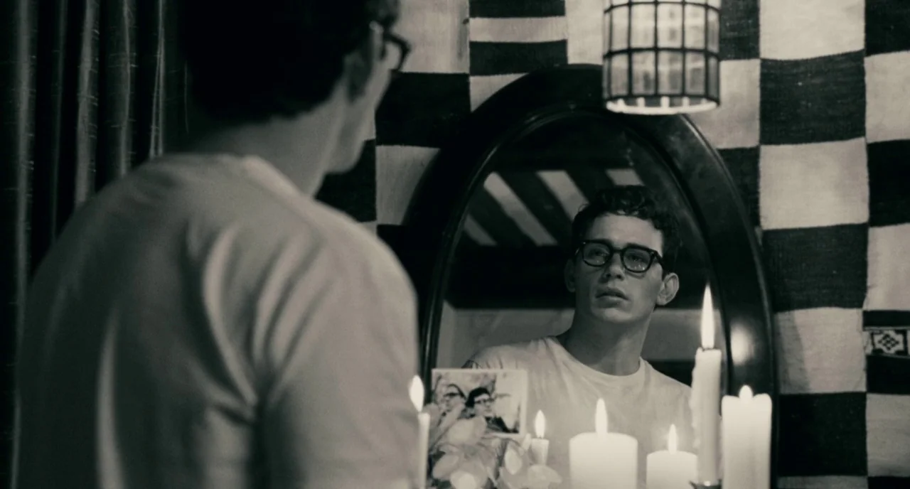 Howl (2010) movie still: medium — A man observes his reflection in an oval mirror mounted on a black-and-white checkered wall. Several lit candles…