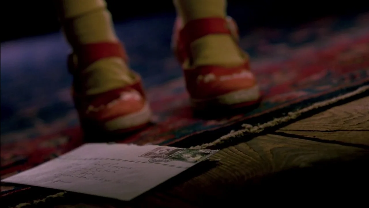 In Dreams (1999) movie still: closeup — An ornate red-patterned rug sits on a wooden floor near a dark space. A white envelope with a stamp lies on…