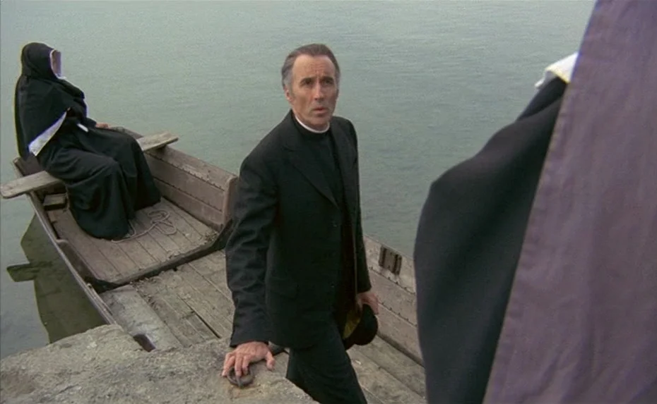 To The Devil a Daughter (1976) movie still: medium — A wooden rowboat sits at a grey stone pier on calm water. A man in a dark suit prepares to…