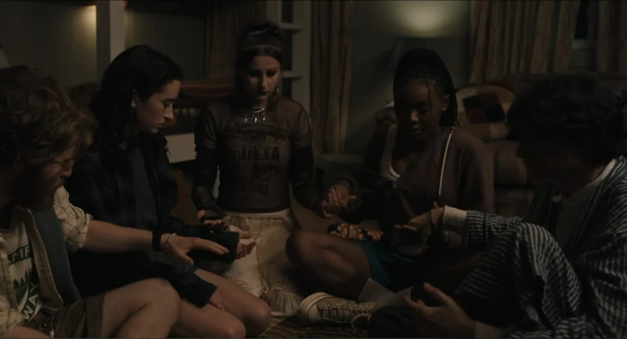 Hell of a Summer (2025) movie still: medium — Five young people sit in a circle on a rug in a dimly lit home. They hold each other's hands while…