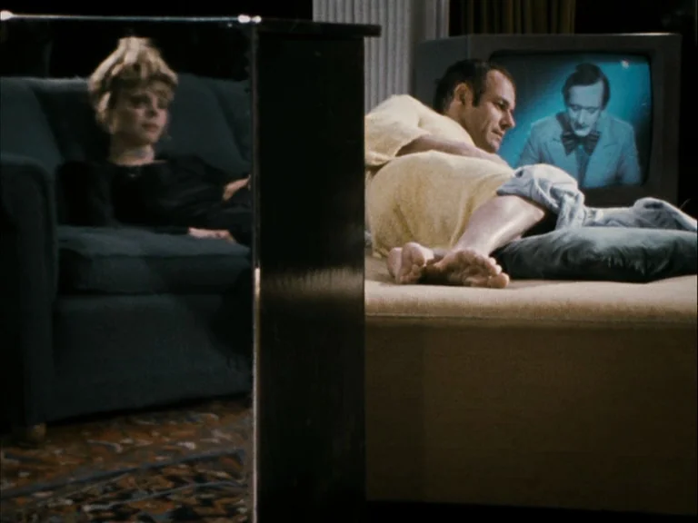 Making of: World On A Wire (1973) movie still: medium — A dimly lit room features a man resting on a sofa watching a television program. A mirror in…