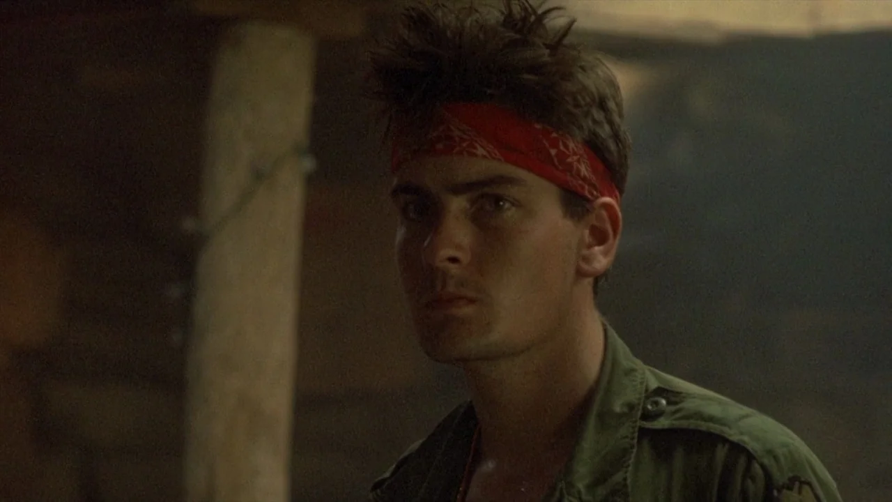 Platoon (1986) movie still: closeup — A dimly lit interior space with canvas walls and wooden support posts visible. A young man with a red headband…