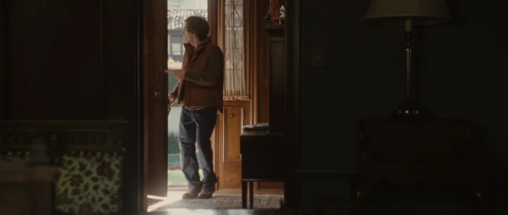 Greenberg (2010) movie still: fullbody — A dark, wood-paneled interior space features an open door leading to an exterior. A man wearing a brown…