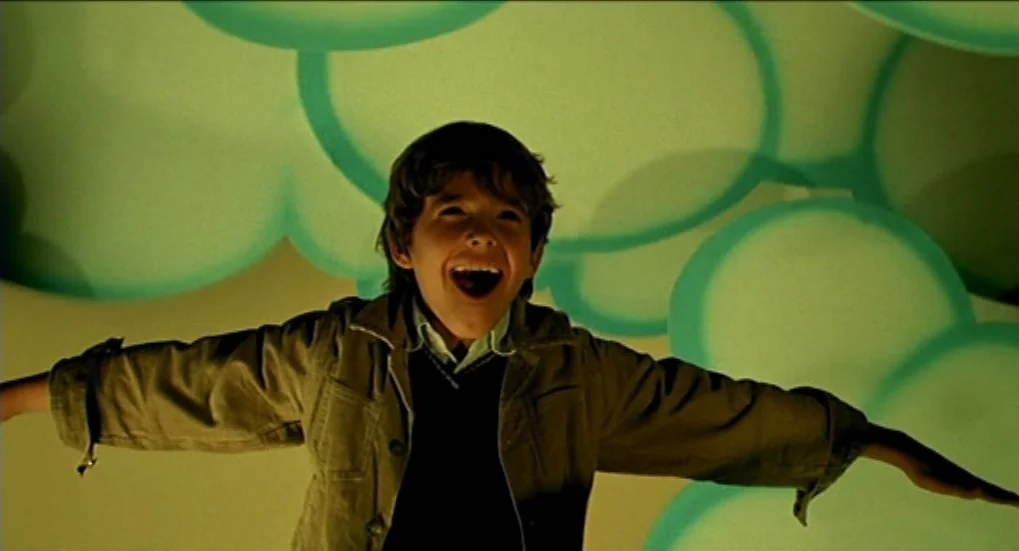 Jeux d’Enfants (2003) movie still: medium — A studio space with a bright green screen projection of cartoon clouds behind the boy. The young boy…