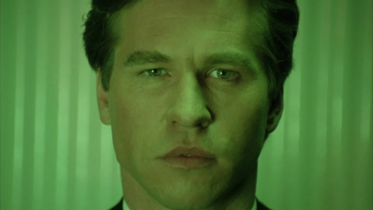 Batman Forever (1995) movie still: closeup — The face of a man is illuminated by intense green light. He stares directly forward against a plain,…