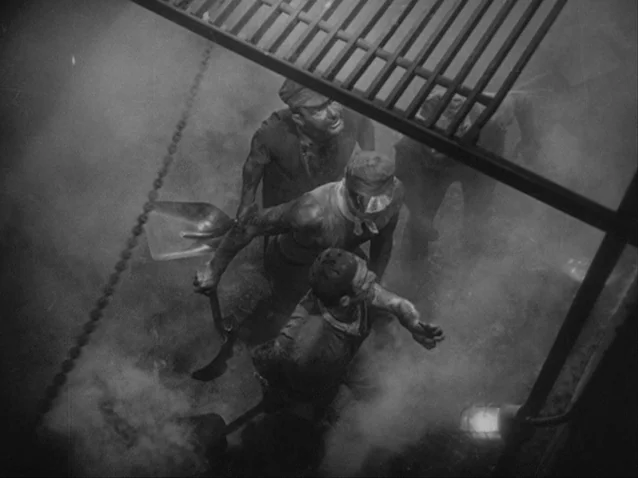 The Docks of New York (1928) movie still: medium — A dark, smoke-filled industrial space features laborers working beneath an overhead metal grate.…