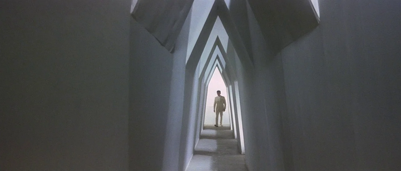 Tokyo Drifter (1966) movie still: wide — A minimalist white interior with sharp, angular archways creating a long tunnel effect. The man walks away…