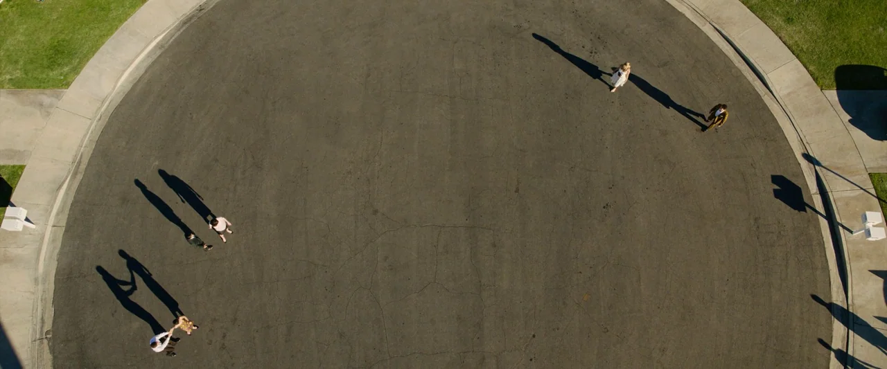 Don’t Worry Darling (2022) movie still: wide — An asphalt cul-de-sac is marked with cracks and bounded by concrete curbs and grass. Four individuals…