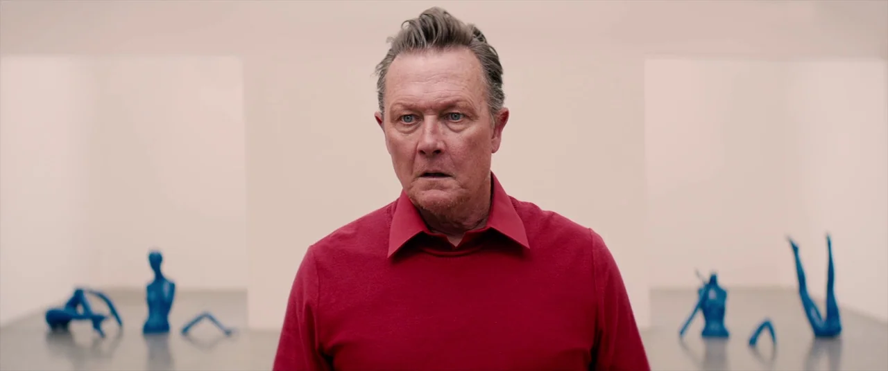 Tone Deaf (2019) movie still: medium — A spacious white art gallery room contains scattered blue mannequin parts on the floor. A man stands in the…