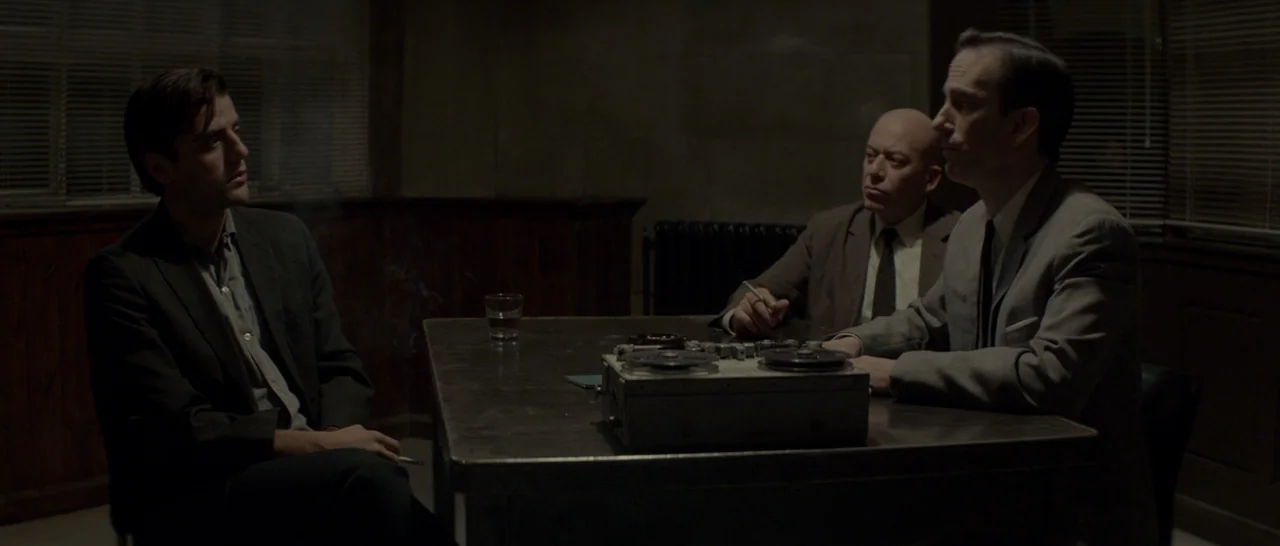 The Two Faces of January (2014) movie still: medium — A dark, spartan room with venetian blinds covering windows on the back wall. Three men sit…