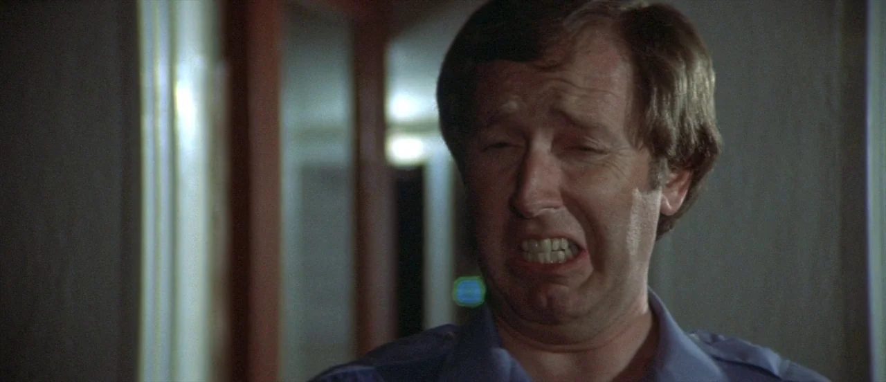 The Long Goodbye (1973) movie still: closeup — A dimly lit indoor hallway with indistinct vertical wall panels. A middle-aged man in a uniform…