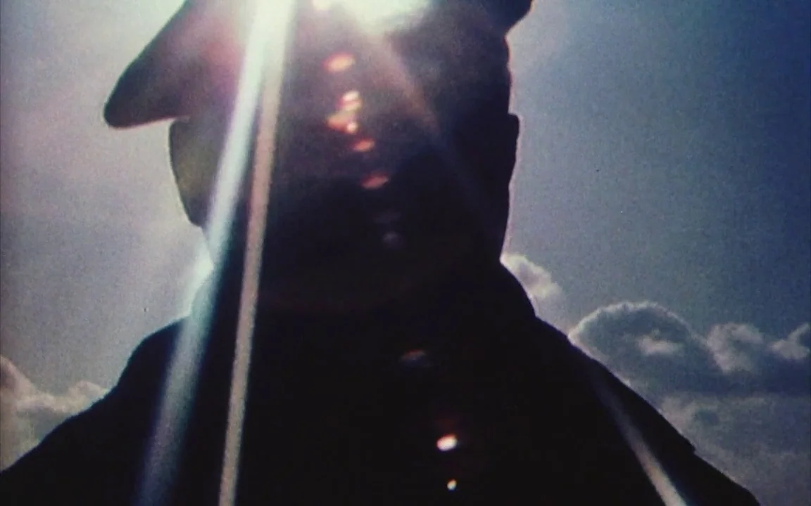 The Last of England (1987) movie still: closeup — The subject stands in an open area against a bright sunlit sky. A dark silhouette of a person…